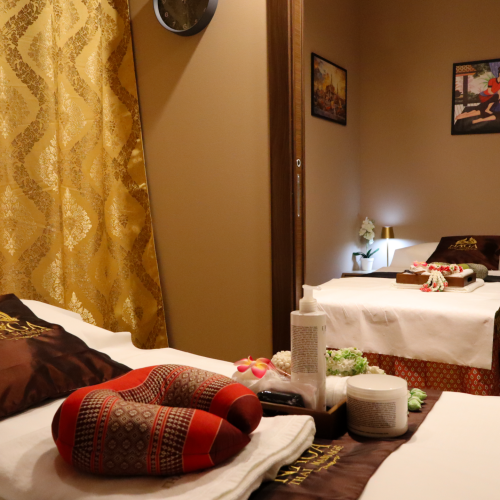 Relaxing Thai spa massage room with two treatment beds, white linens, herbal/beauty products, and warm lighting.